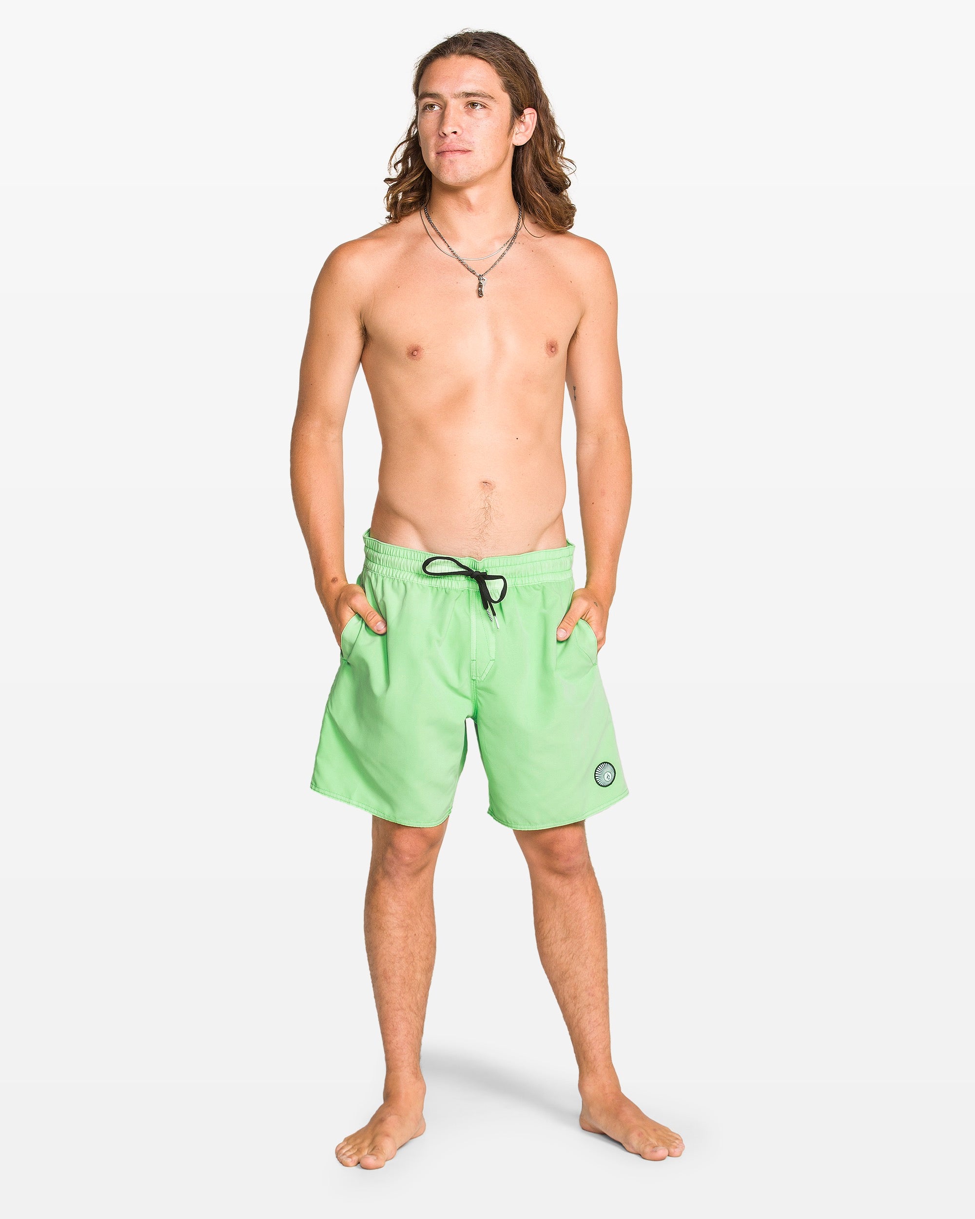 A young man with long brown hair stands barefoot against a white background, hands in pockets, wearing Volcom Men's Center Elastic Waist 17" Trunk swim shorts in light green with black drawstring and circular logo, paired with a necklace.