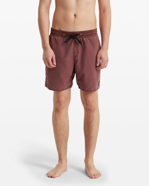 A person stands barefoot, facing forward against a white background, wearing Volcom Men's Center Elastic Waist Trunks in maroon with a black drawstring, elastic waist, and a small patch on the left leg.