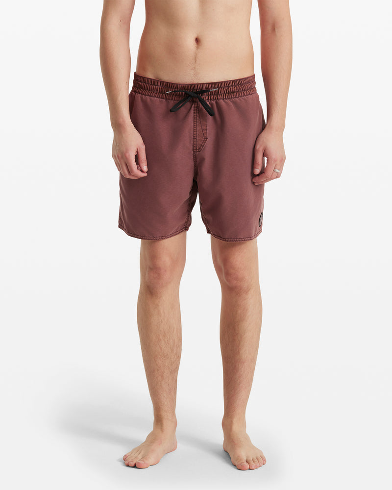 A person stands barefoot, facing forward against a white background, wearing Volcom Men's Center Elastic Waist Trunks in maroon with a black drawstring, elastic waist, and a small patch on the left leg.