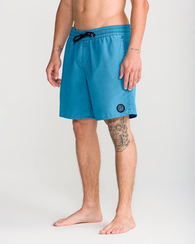 A person stands barefoot wearing Volcom Men’s Center Trunk 17 swim shorts in teal, featuring a black drawstring and a circular Volcom logo near the hem. Designed with a 17-inch outseam for modern style, these stretch trunks reveal tattoos on the wearer’s left forearm and right thigh, while their torso remains partially out of view.