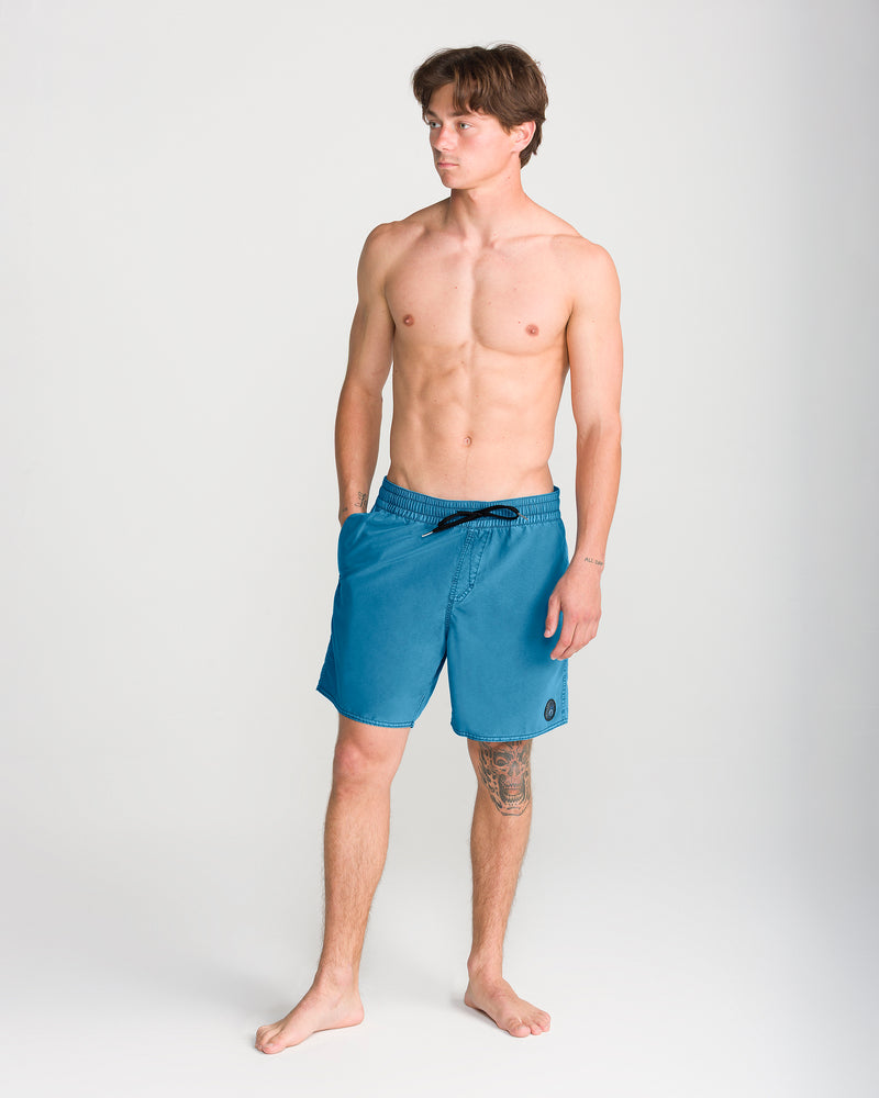 A shirtless man stands barefoot on a white background, wearing Volcom Men’s Center Trunk 17 swim trunks in blue. He has short brown hair, a tattoo on his left thigh, and is looking slightly to his left.