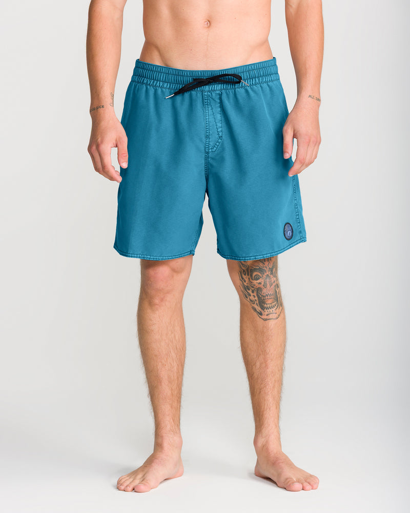 A person standing barefoot, wearing Volcom Men’s Center Trunk 17 swim shorts in teal, featuring a black drawstring and a circular logo on the left leg. These men’s swim trunks have a 17-inch outseam, and the individual has a tattoo on their right shin.