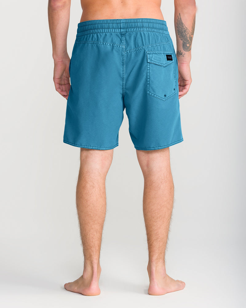 A person stands barefoot facing away, wearing Volcom Men’s Center Trunk 17 swim trunks in teal, featuring a 17-inch outseam, a back pocket, and a small sewn label. A tattoo is visible on their left arm against a plain light-colored background.