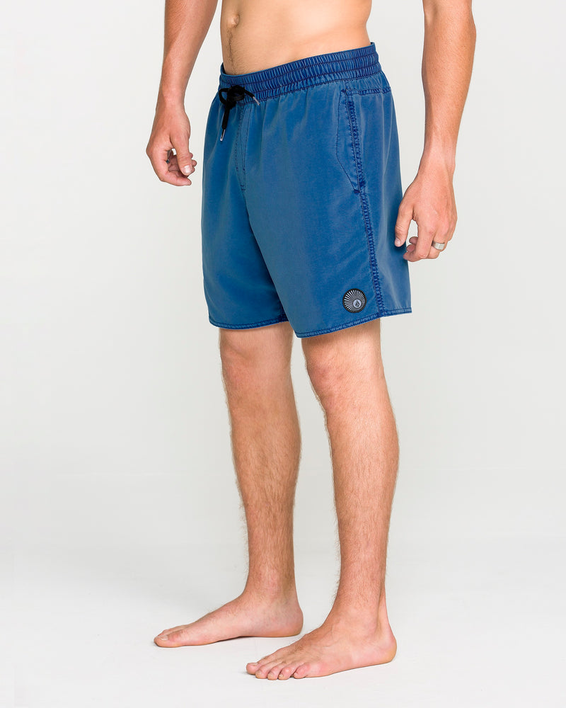 A person stands barefoot on a light background, wearing Volcom Men's Center Elastic Waist Trunks in blue with a 17-inch length, featuring a black drawstring and a circular patch on the left leg. The upper body is not visible, drawing attention to the stylish swimwear.