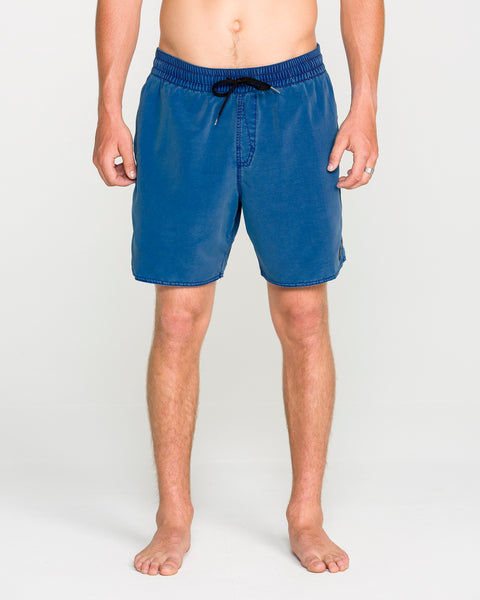 A person standing barefoot, wearing Volcom Men's Center Elastic Waist Trunks in blue with a black drawstring—classic men's swimwear—shown from shoulders to feet against a plain light background.