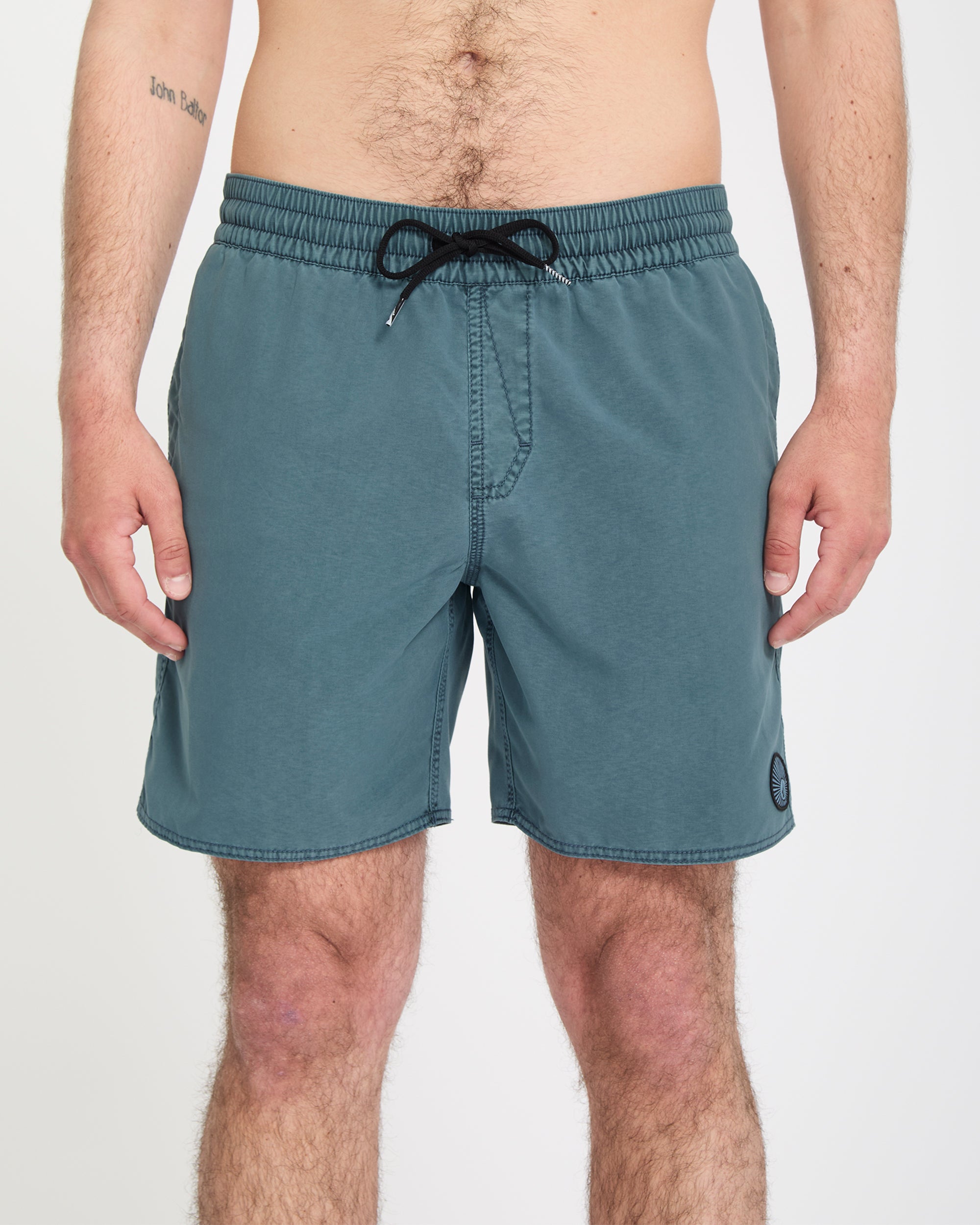 A person stands facing forward in Volcom Men's Center Trunks 17, teal swim shorts with side pockets and a small patch logo on the front left leg. Their arms rest at their sides against a plain white background.