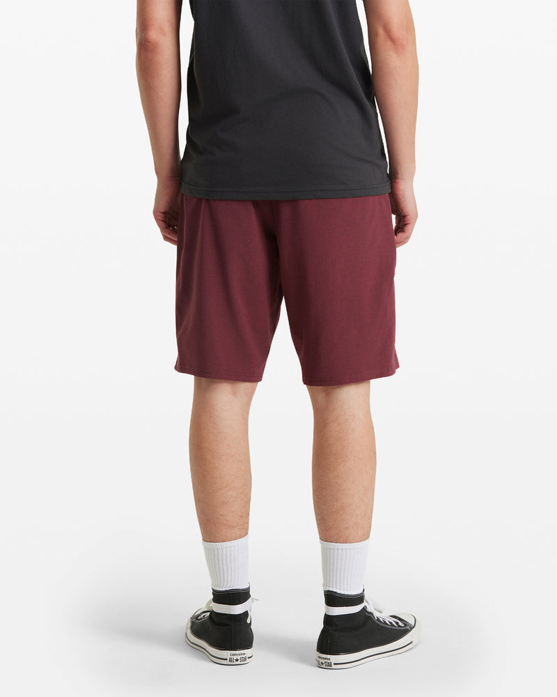 A person stands facing away in a black t-shirt, Men's Frickin Cross Shred Static Shorts by Volcom in maroon, white socks, and black low-top sneakers against a plain white background.
