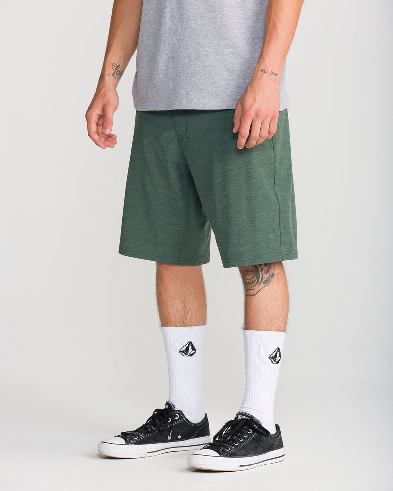 A person wearing a gray t-shirt, Volcom Men’s Frickin Cross Shred Static 20 modern-fit shorts in green made from recycled polyester, white socks featuring black logos, and black sneakers. The individual has tattoos on their left forearm and right leg. The background is plain and light-colored.