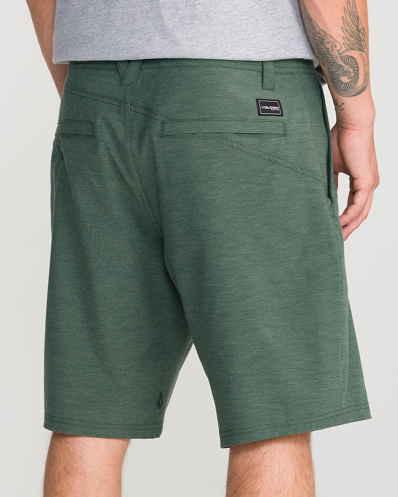 A person wearing Volcom Men’s Frickin Cross Shred Static 20 shorts, crafted from recycled polyester, and a gray shirt stands with their left hand in their pocket, displaying a tattoo on their right arm. The Volcom logo patch is visible on the back pocket of these modern-fit shorts.