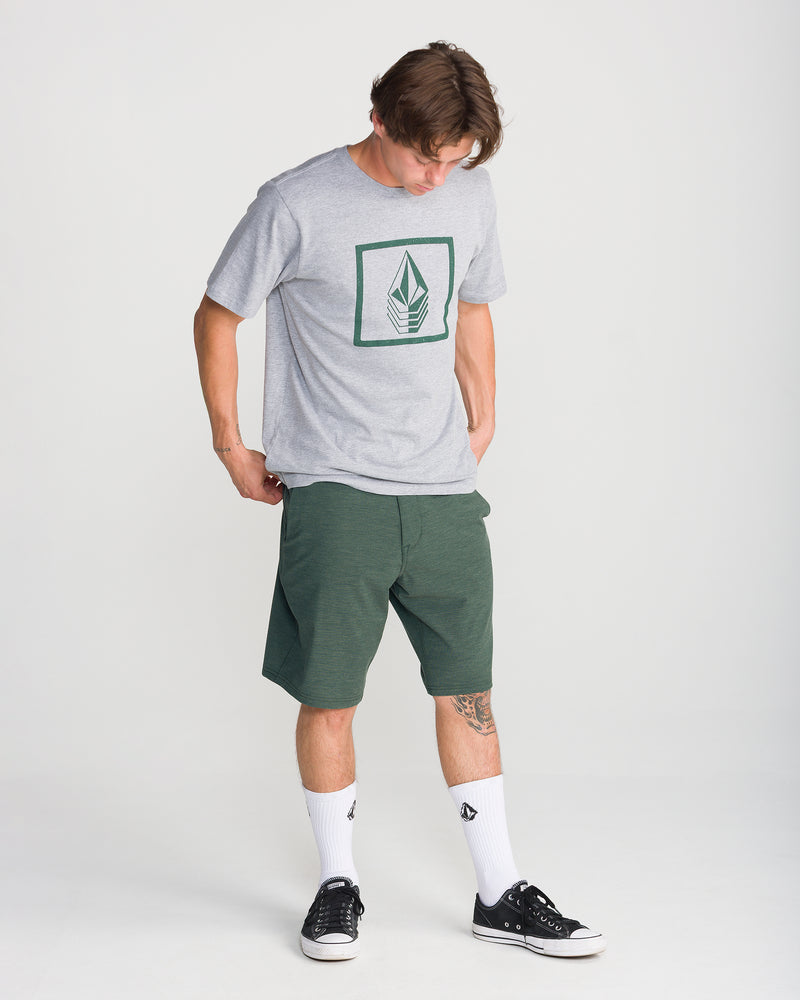 A person in a gray graphic t-shirt, white crew socks, and black sneakers stands looking down on a plain white background while wearing modern-fit green Volcom Men’s Frickin Cross Shred Static 20 shorts crafted from recycled polyester.