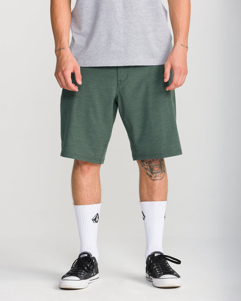 A person wearing a grey T-shirt, Volcom Men’s Frickin Cross Shred Static 20 modern-fit green shorts that fall to the knees, white socks with a black logo, and black sneakers. The image is cropped from shoulders down; one sneaker lace is untied.