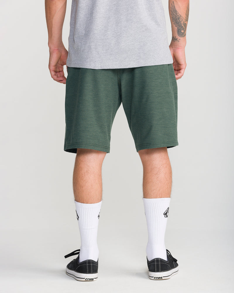A person stands facing away, wearing a gray t-shirt, the Volcom Men’s Frickin Cross Shred Static 20 green shorts made from recycled polyester, white crew socks, and black-and-white sneakers. Part of a tattoo is visible on their left arm. The background is plain and light.
