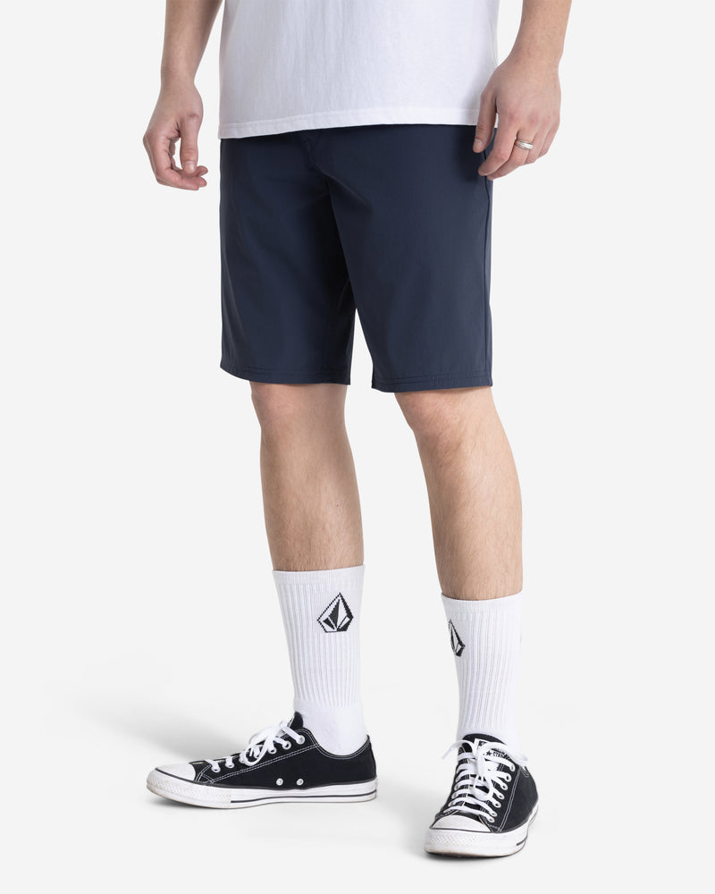 The lower half of a person is shown against a plain white background, wearing Volcom Men’s Frickin Cross Shred Shorts with a 20" outseam, a white t-shirt, white logo socks, and black low-top sneakers.