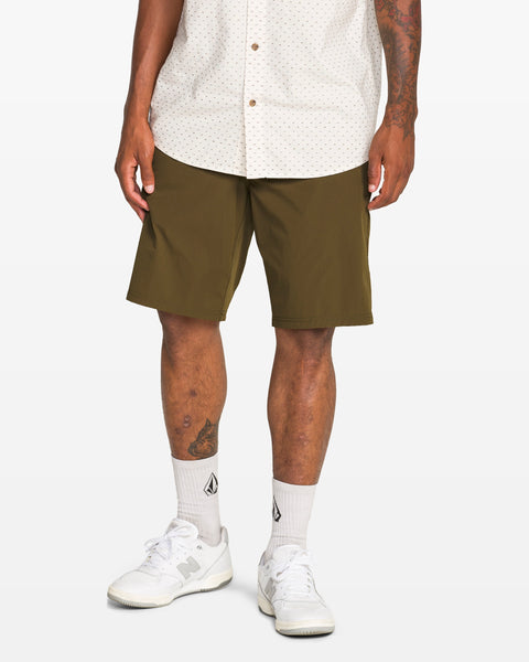 A person stands against a plain white background wearing a white patterned short-sleeve shirt, Volcom Men's Frickin Cross Shred Shorts in olive green (20" outseam), white socks with a black logo, and white New Balance sneakers.
