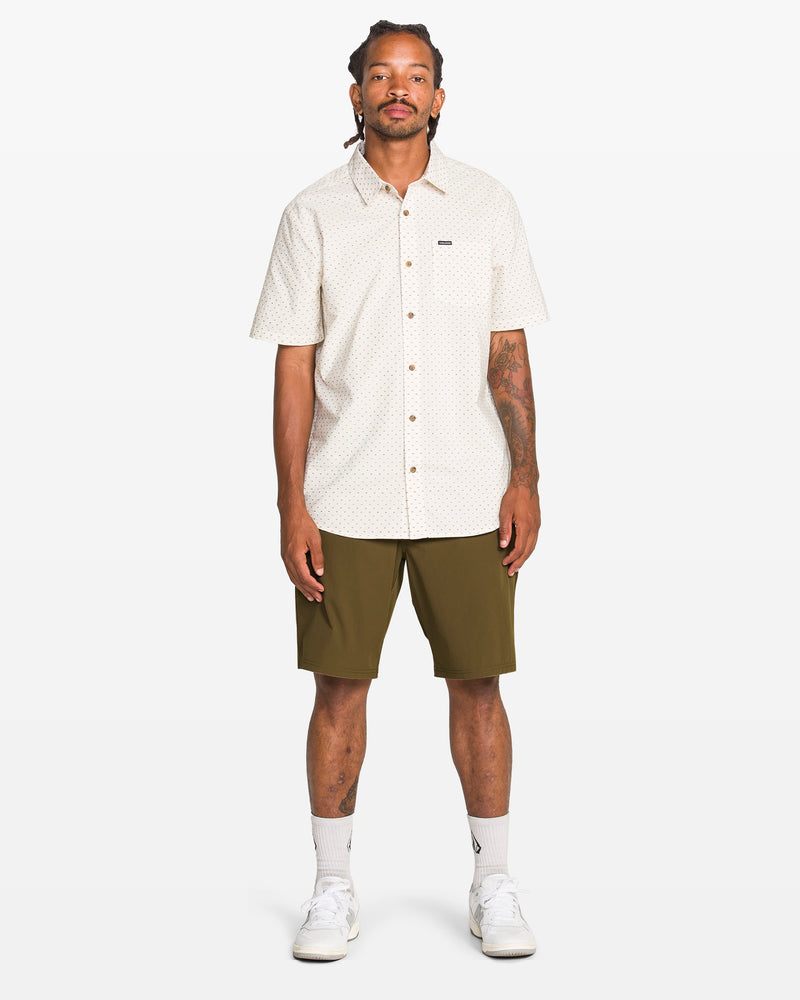 A person stands facing forward against a plain white background, wearing a light-colored short-sleeve button-up shirt, Volcom Men's Frickin Cross Shred Shorts in olive green, white socks, and white sneakers.