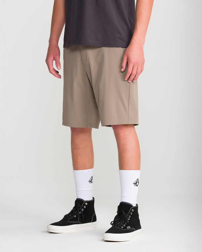 A person wearing the Volcom Men’s Frickin Cross Shred 20 beige knee-length shorts, a dark t-shirt, white crew socks with a black logo, and black high-top sneakers stands against a plain white background.
