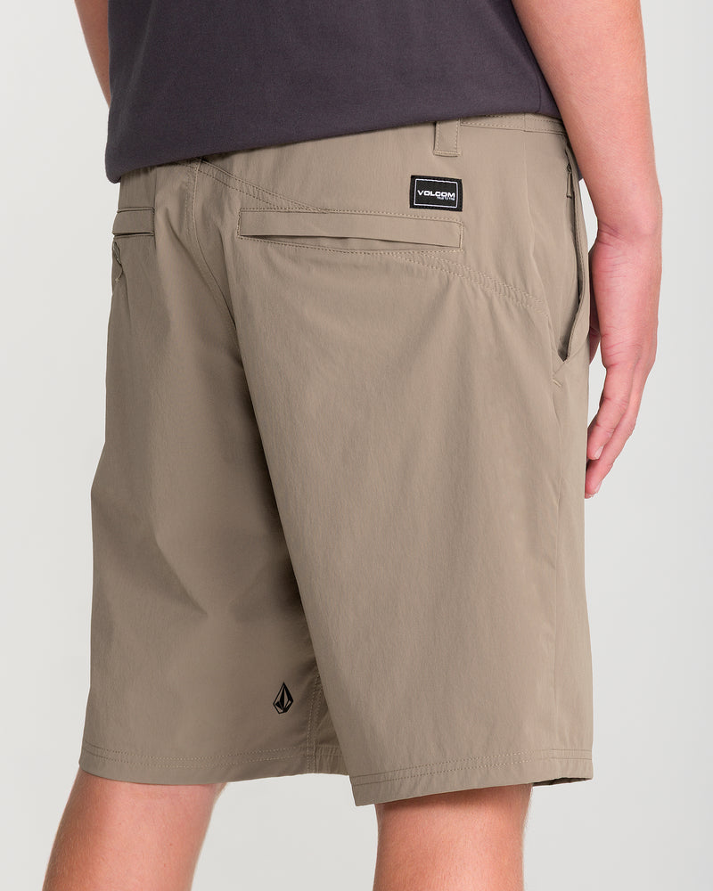 A person wearing Volcom Men’s Frickin Cross Shred 20 water-repellent shorts in light brown and a dark shirt stands facing away, highlighting the back and side view of the stretch fabric shorts with a Volcom logo patch above the pocket.