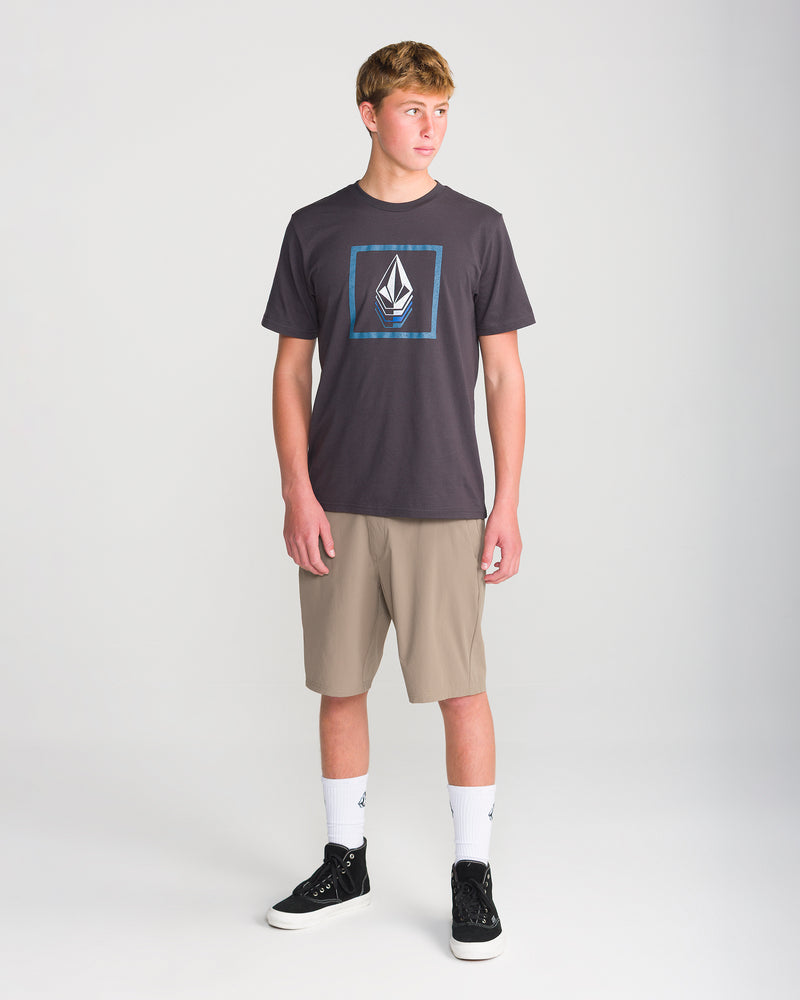 A young person stands in a studio against a plain light background, looking slightly to their left and wearing a dark graphic t-shirt, white socks, black sneakers, and Volcom’s Men’s Frickin Cross Shred 20 tan knee-length stretch fabric shorts with antimicrobial treatment.