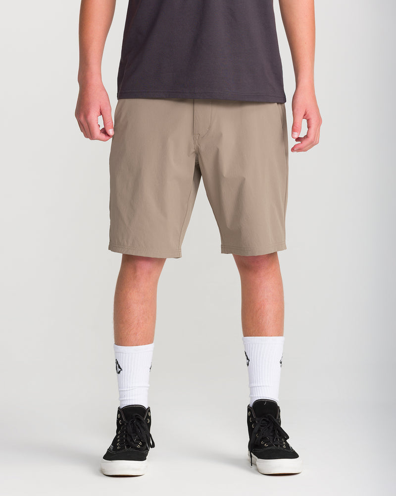 A person wearing a dark t-shirt, Volcom Men’s Frickin Cross Shred 20 beige knee-length water repellent shorts, white socks with black logos, and black sneakers stands against a plain background.