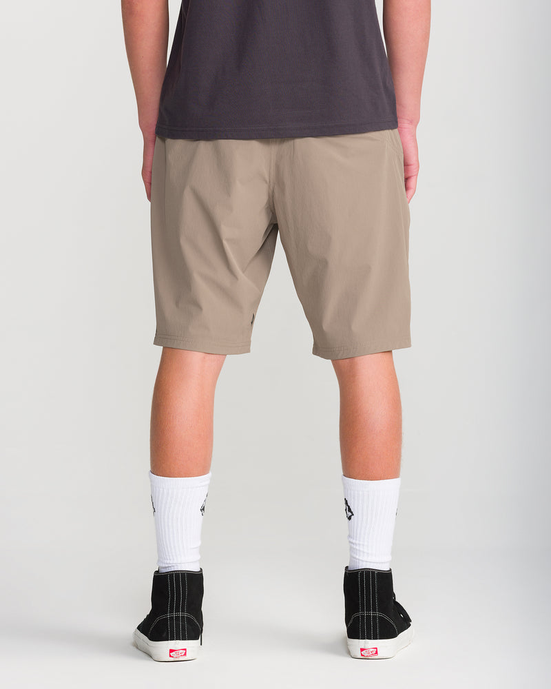 A person stands facing away, wearing a black shirt, white socks, black Vans sneakers, and tan knee-length stretch fabric shorts—the Volcom Men’s Frickin Cross Shred 20. The background is plain and light-colored.