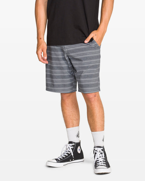 A person stands with one hand in their pocket against a plain white background, wearing a black t-shirt, Volcom Men's Mix Frickin Cross Shred Shorts in gray stripe with fixed waist, white socks with a black logo, and black high-top sneakers.