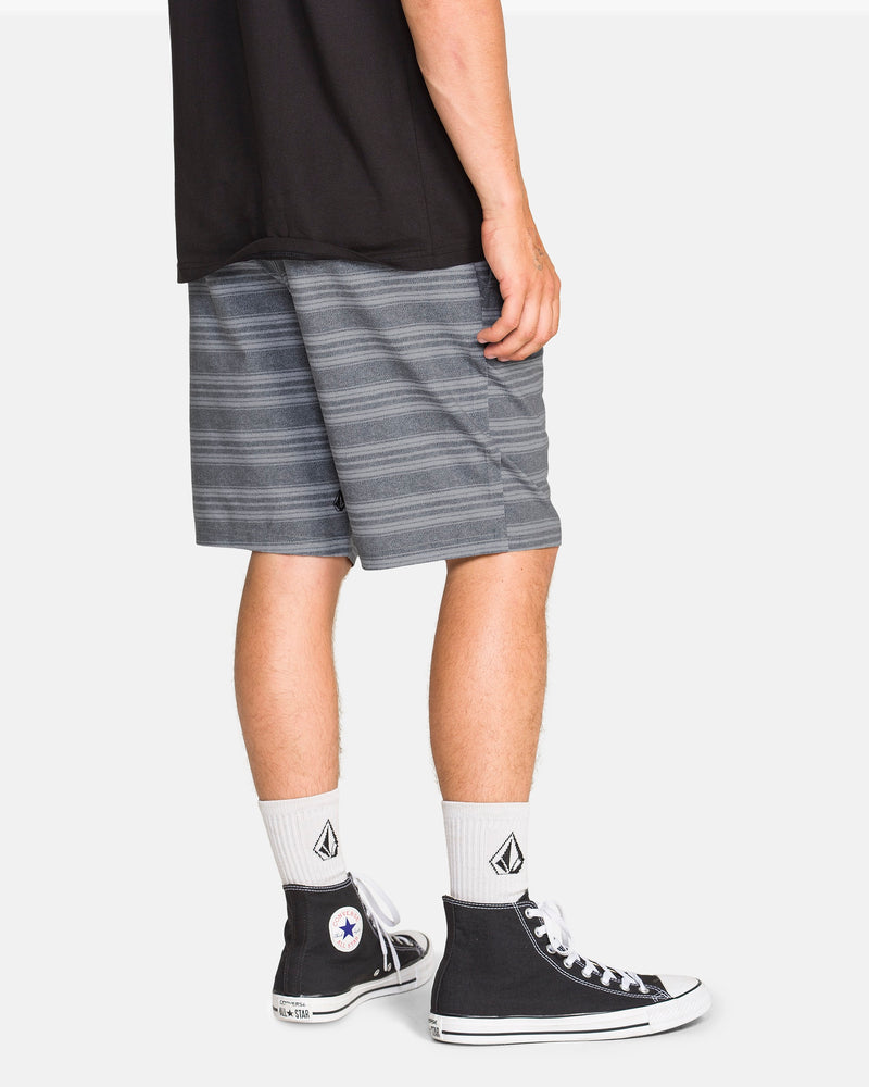 A person wearing a black T-shirt, Volcom Men's Mix Frickin Cross Shred Shorts, white socks with a black logo, and black high-top Converse sneakers stands against a plain white background.