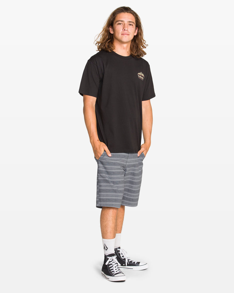 A young man with long brown hair stands against a white background, wearing Volcom Men's Mix Frickin Cross Shred Shorts, a black T-shirt, white socks, and black high-top sneakers, smiling with his hands relaxed by his sides.