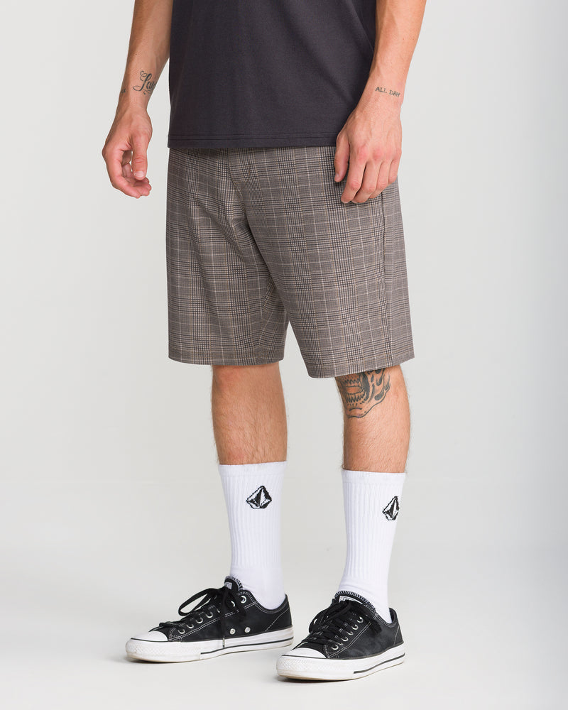 A person wearing Volcom Men’s Mix Frickin Cross Shred 20 shorts in a Desert Palm plaid pattern, paired with a dark t-shirt, white socks featuring a black logo, and black sneakers stands against a plain background. Tattoos are visible on their arm and leg.
