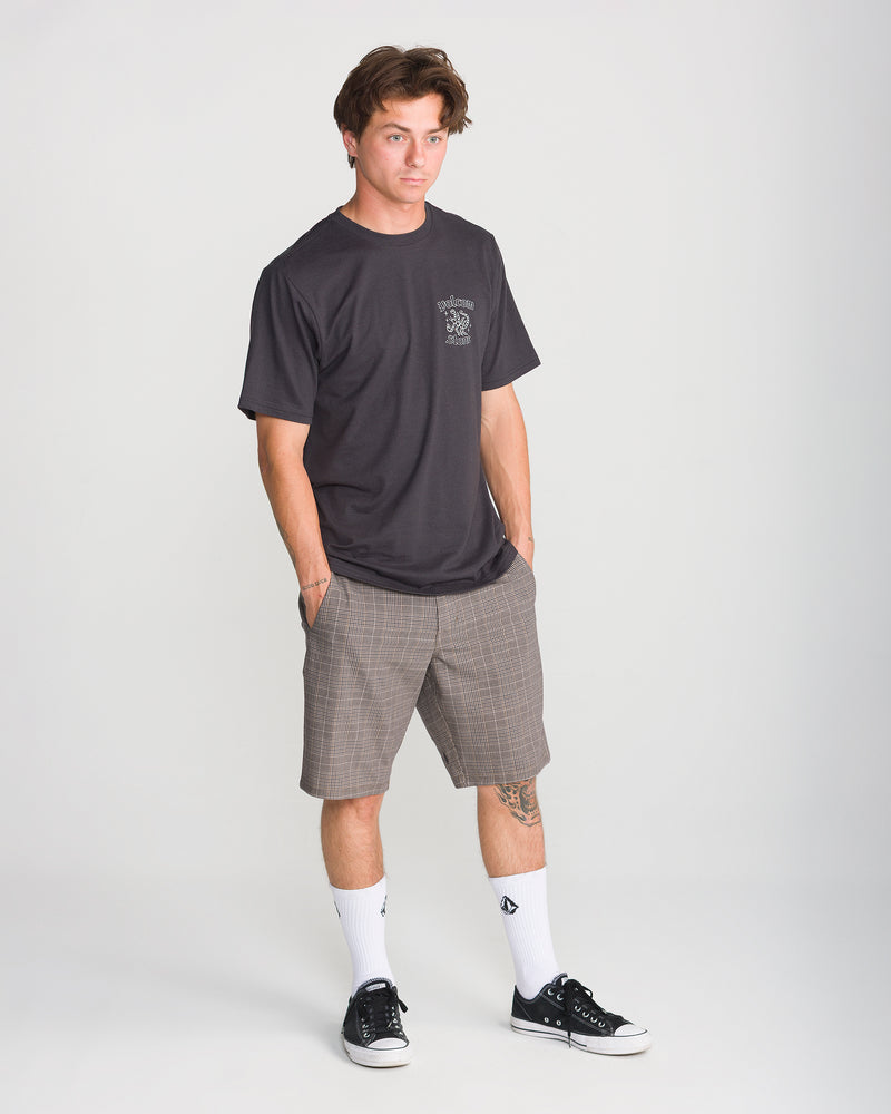 A person wearing a black t-shirt, Volcom Men’s Mix Frickin Cross Shred 20 shorts, white socks, and black sneakers stands against a plain light background with hands in pockets, looking slightly to the side.