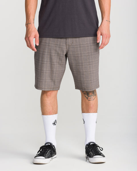 A person wearing Volcom Men’s Mix Frickin Cross Shred 20 shorts in the Desert Palm plaid pattern, paired with a dark shirt, white socks featuring a black logo, and black sneakers stands against a plain background. Only the lower half of their body is visible.