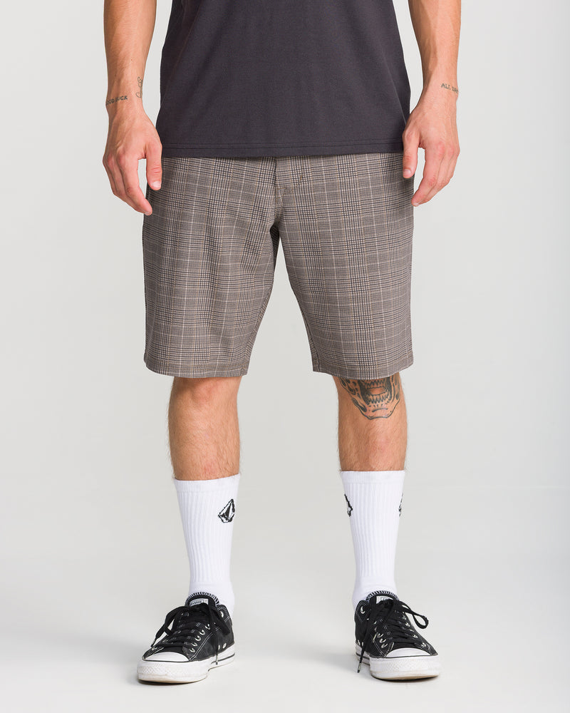 A person wearing Volcom Men’s Mix Frickin Cross Shred 20 shorts in the Desert Palm plaid pattern, paired with a dark shirt, white socks featuring a black logo, and black sneakers stands against a plain background. Only the lower half of their body is visible.