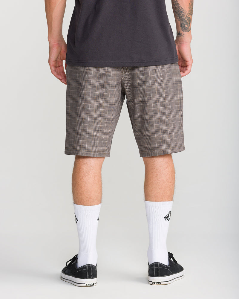 A person stands facing away, wearing a dark shirt and the Volcom Men’s Mix Frickin Cross Shred 20 boardshorts in Desert Palm plaid, which fall to the knees. White crew socks with logos and black sneakers complete the look, with part of a tattoo visible on their left arm. The background is plain light gray.
