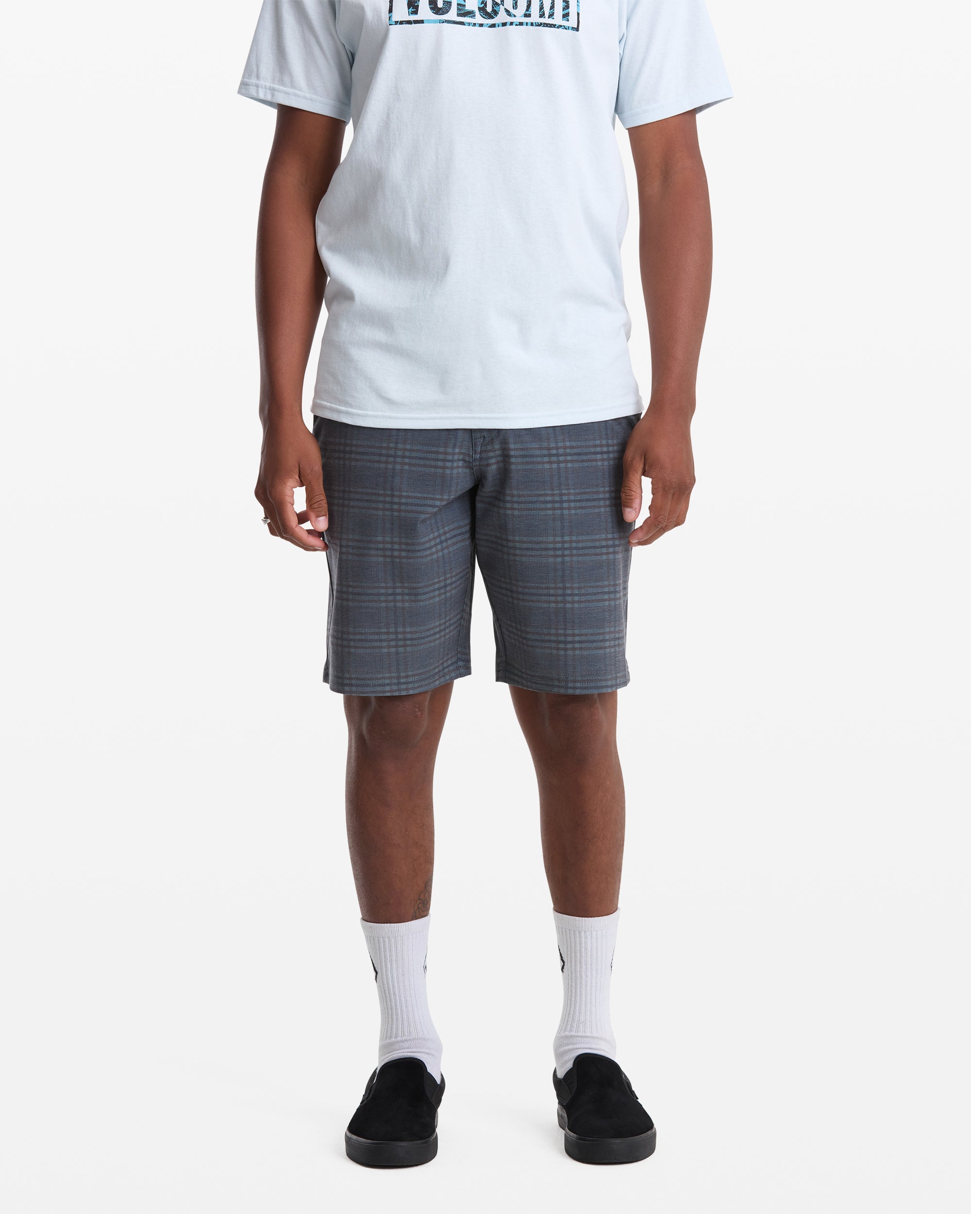 A person stands facing forward against a white background, wearing a light blue T-shirt, white socks, black slip-on shoes, and Volcom Men's Mix Frickin Cross Shred Shorts. Their face is not visible.
