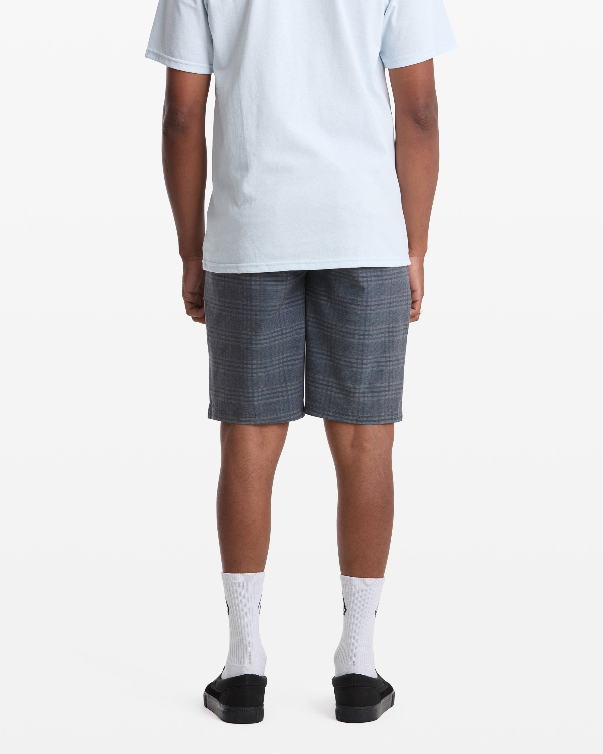 A person stands facing away against a plain white background, wearing Volcom Men's Mix Frickin Cross Shred Shorts in gray plaid, a light blue T-shirt, white crew socks, and black slide sandals.