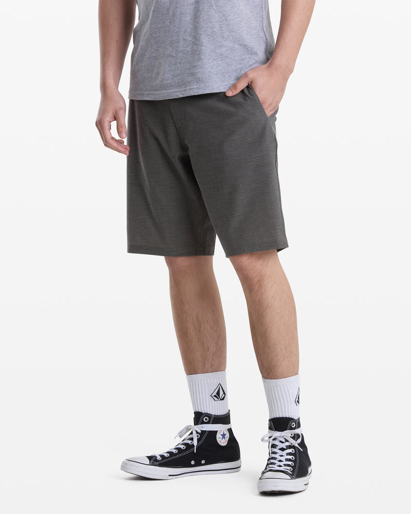 A person wears a gray t-shirt, Volcom Men's Mix Frickin Cross Shred Shorts in dark gray, white crew socks with a black logo, and black Converse high-top sneakers, standing with one hand in their pocket against a plain white background.