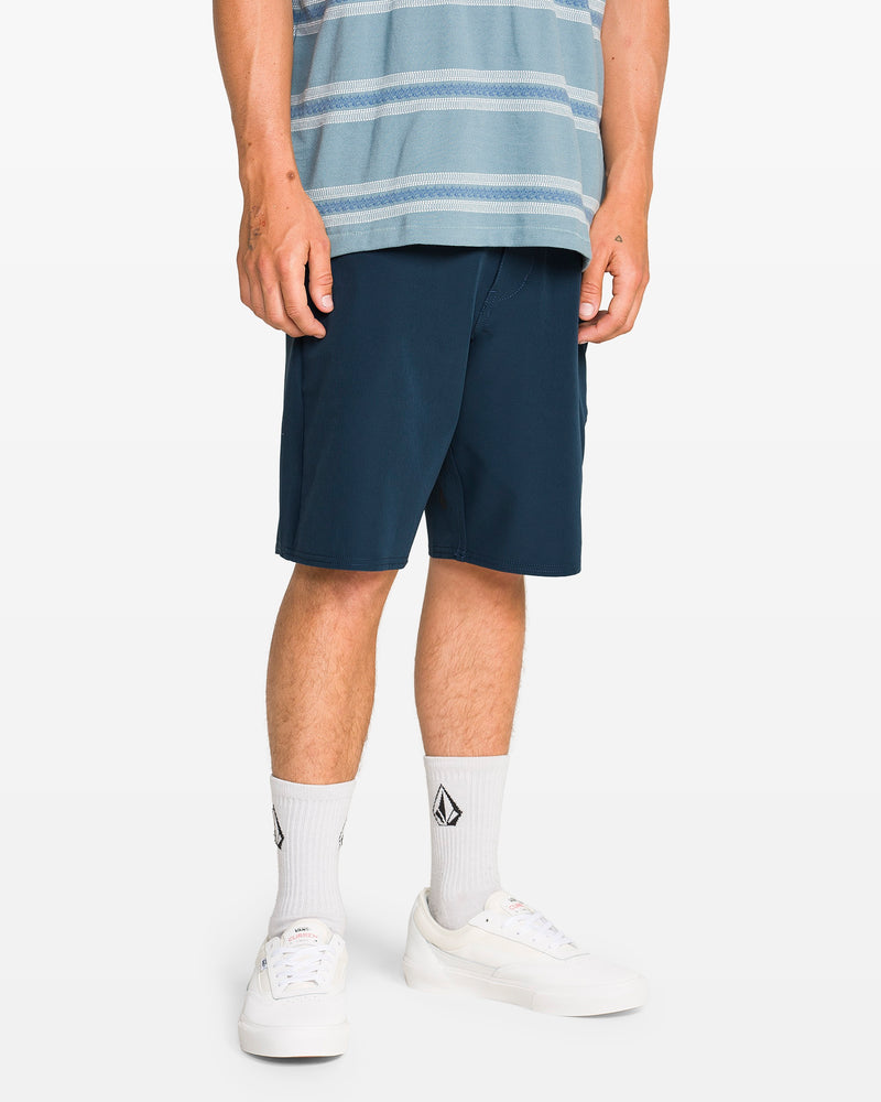 A person wearing a blue striped shirt, Volcom Men's Harbor Hybrid Shorts with a 20-inch outseam, white socks with a black logo, and white sneakers stands against a plain white background.
