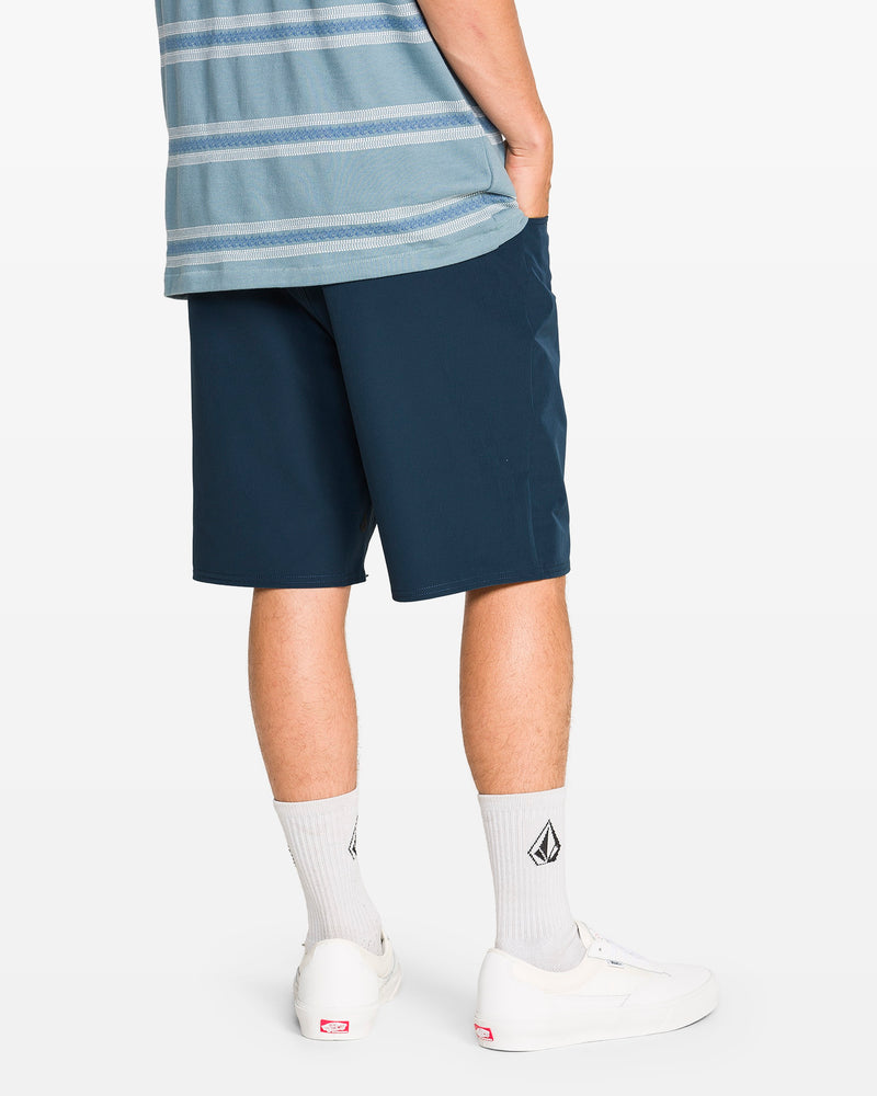 A person, shown from the back against a plain background, wears a blue and gray striped shirt with Volcom Men's Harbor Hybrid Shorts in navy (20-inch outseam), paired with white logo socks and white sneakers, one hand in their pocket.