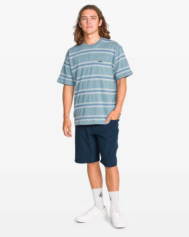 A young man with long hair wears a blue striped tee, Volcom Men's Harbor Hybrid Shorts, white socks, and sneakers while standing against a plain white background.