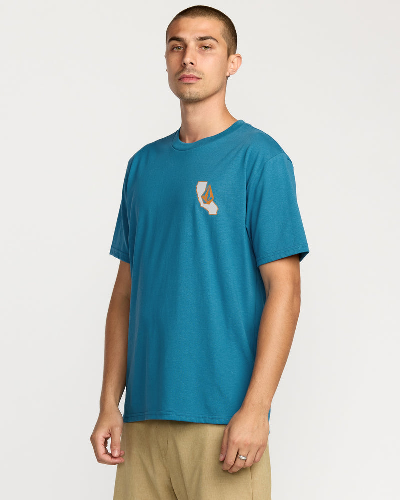 A man wears the Volcom Men's California Dreaming Short Sleeve Tee in teal, featuring a soft-hand screen print on the chest, paired with beige pants, standing against a plain white background.