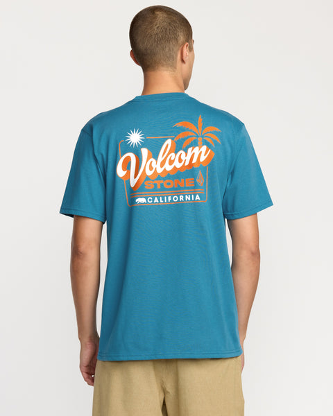 A person, seen from behind, is wearing the Volcom Men's California Dreaming Short Sleeve Tee in blue, which features an orange and white palm tree, sun, and "California" graphic on the back.