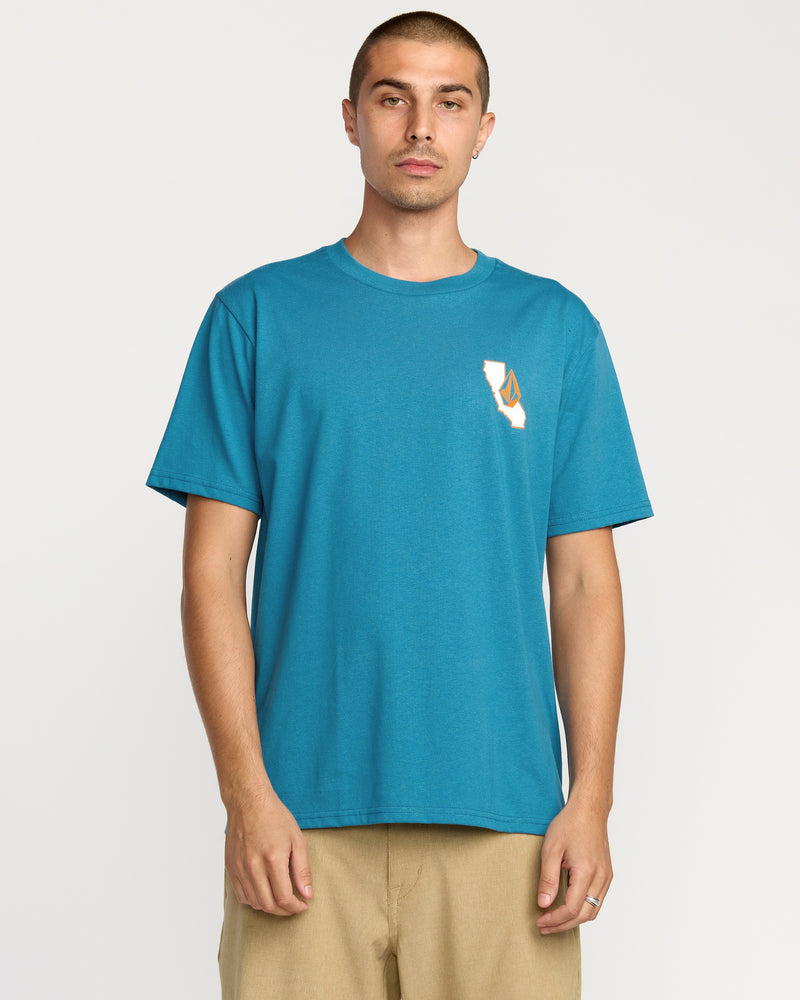 A man with short hair wears the Volcom Men's California Dreaming Short Sleeve Tee in teal and beige pants, standing against a white background. The classic fit tee features a small soft-hand screen print graphic on the left chest.