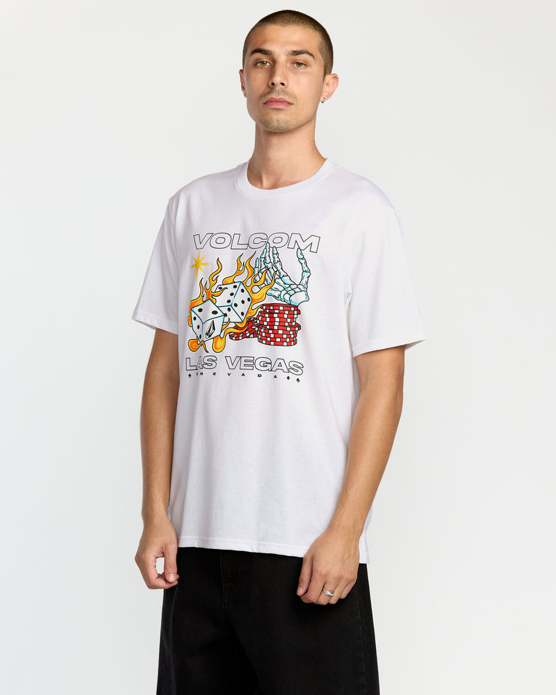 A man wears the Volcom Men's Pass Line Short Sleeve Tee, a white crew T-shirt with dice, flames, cards, and "VOLCOM LAS VEGAS" graphic. He faces forward with a neutral look and black pants.