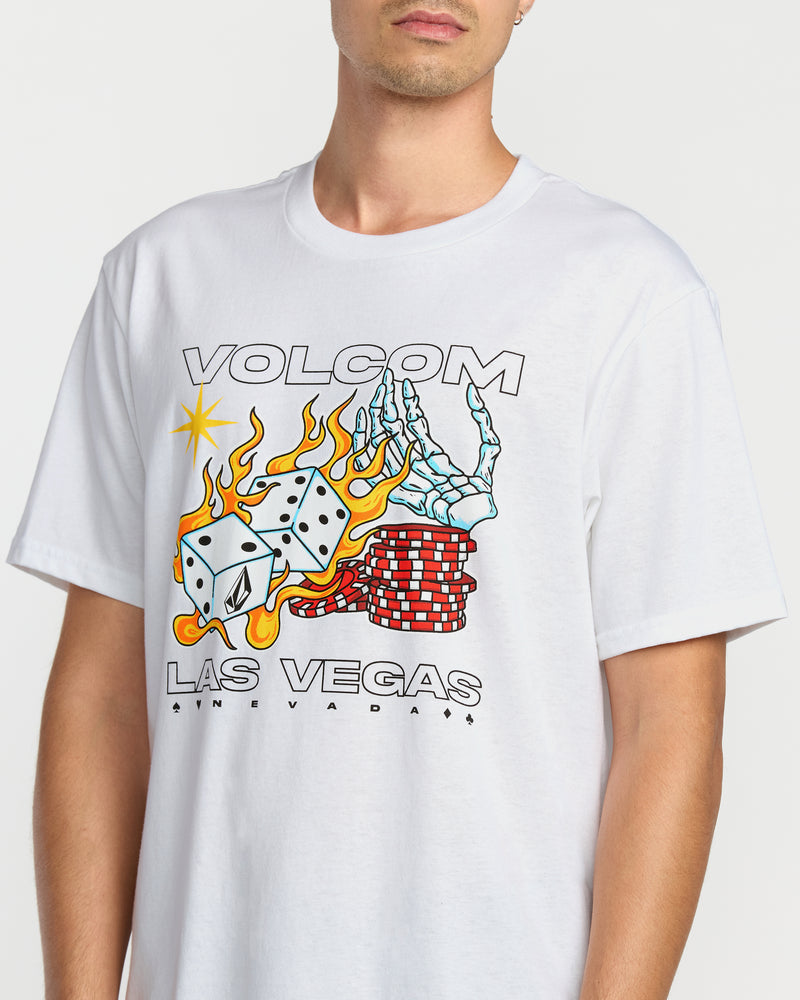 A person wearing the Volcom Men's Pass Line Short Sleeve Tee, a classic fit white crewneck featuring a graphic of a skeleton hand, burning dice, poker chips, and "VOLCOM LAS VEGAS SIN CITY" on the front.