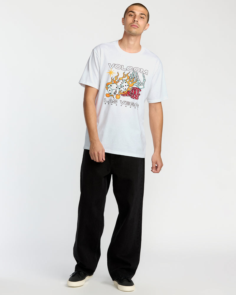 A man stands against a white background wearing the Volcom Men's Pass Line Short Sleeve Tee featuring colorful Las Vegas graphics and “VOLCOM LAS VEGAS” text, paired with loose black pants and black sneakers.