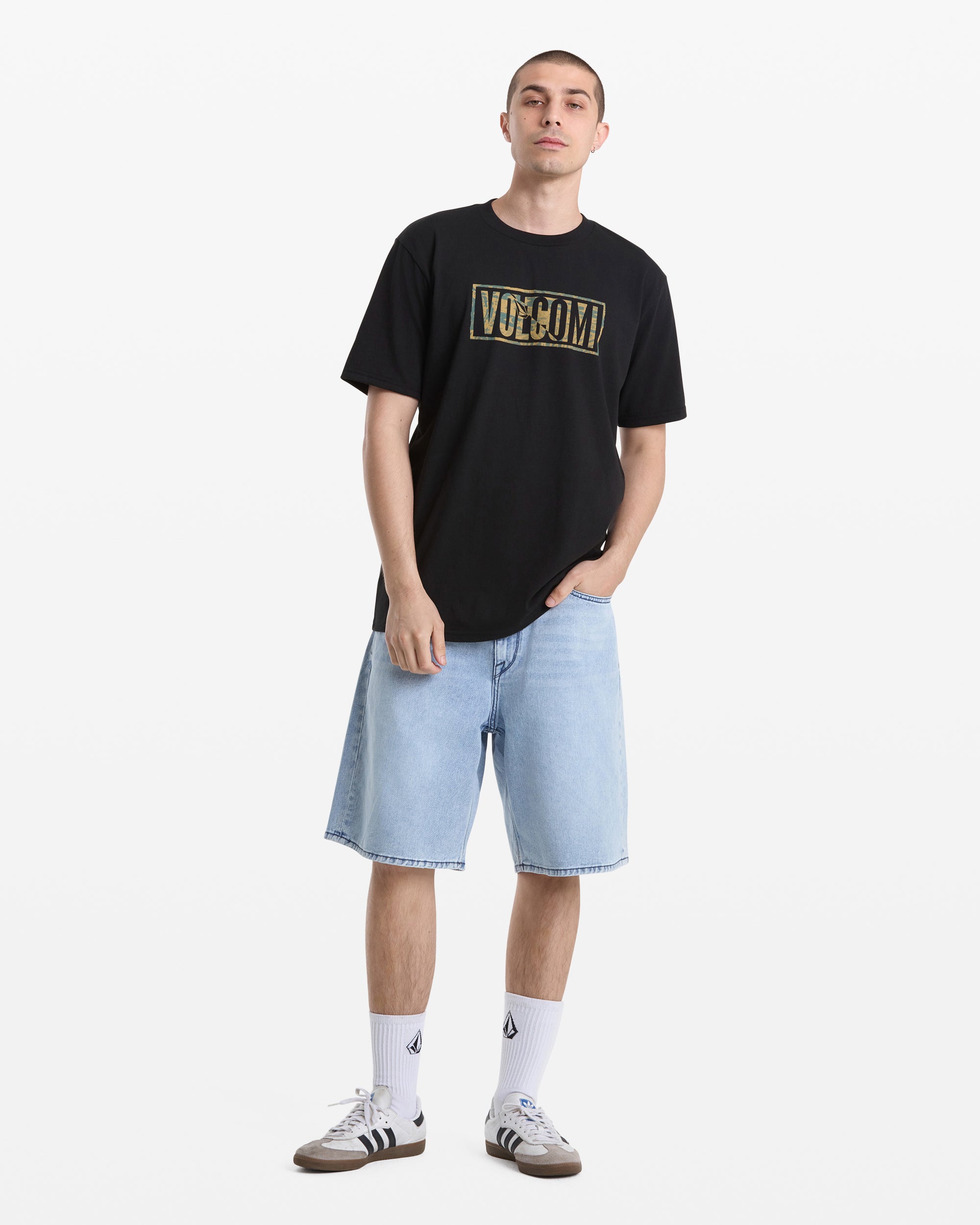 A young man stands against a white background wearing a Volcom Men's Chaotic Short Sleeve Tee in black, light blue knee-length denim shorts, white socks with black logos, and brown sneakers. One hand is in his pocket.