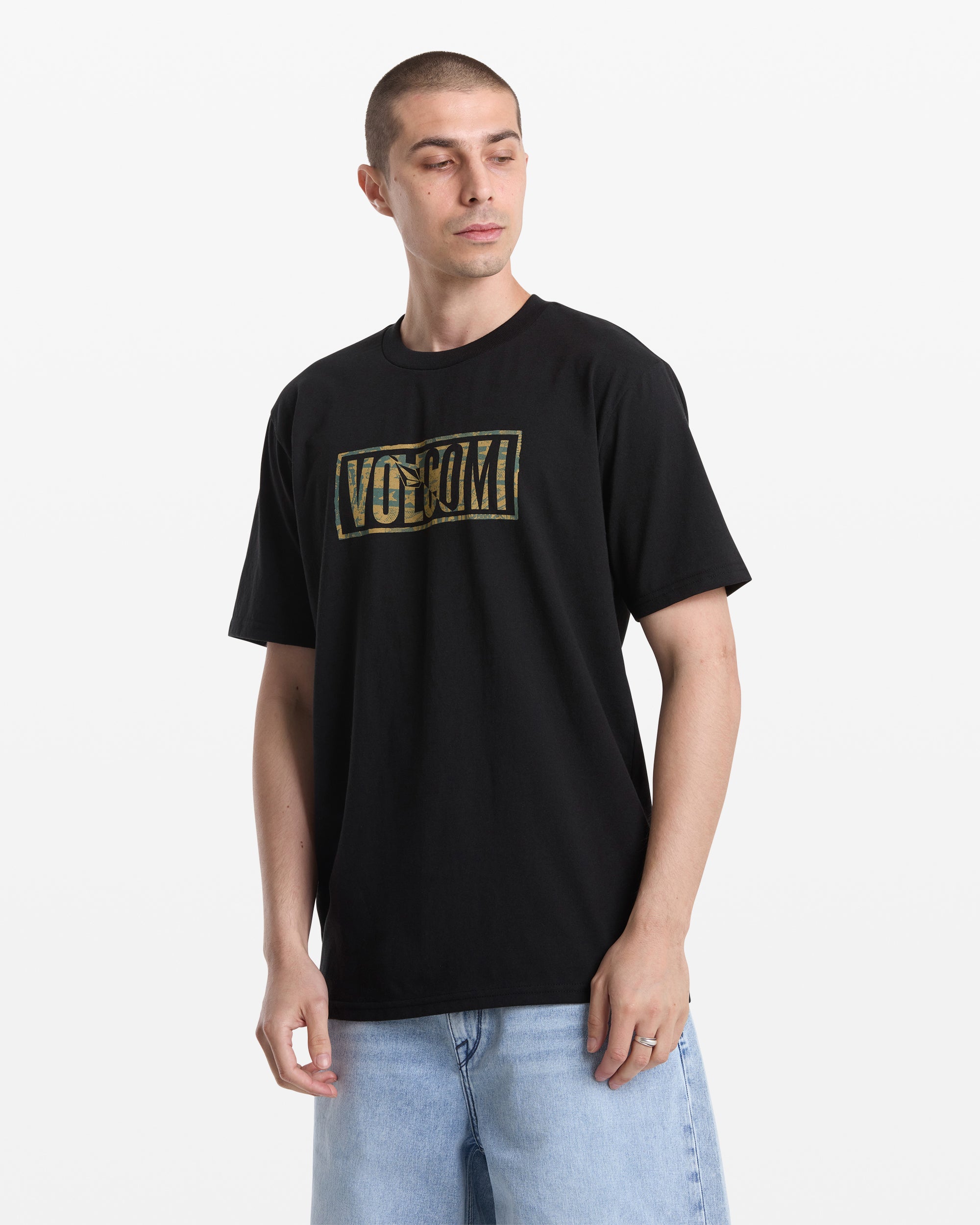 A man with short hair wears the Volcom Men's Chaotic Short Sleeve Tee in black, featuring a gold logo on the chest, paired with light blue jeans, and stands against a plain white background.