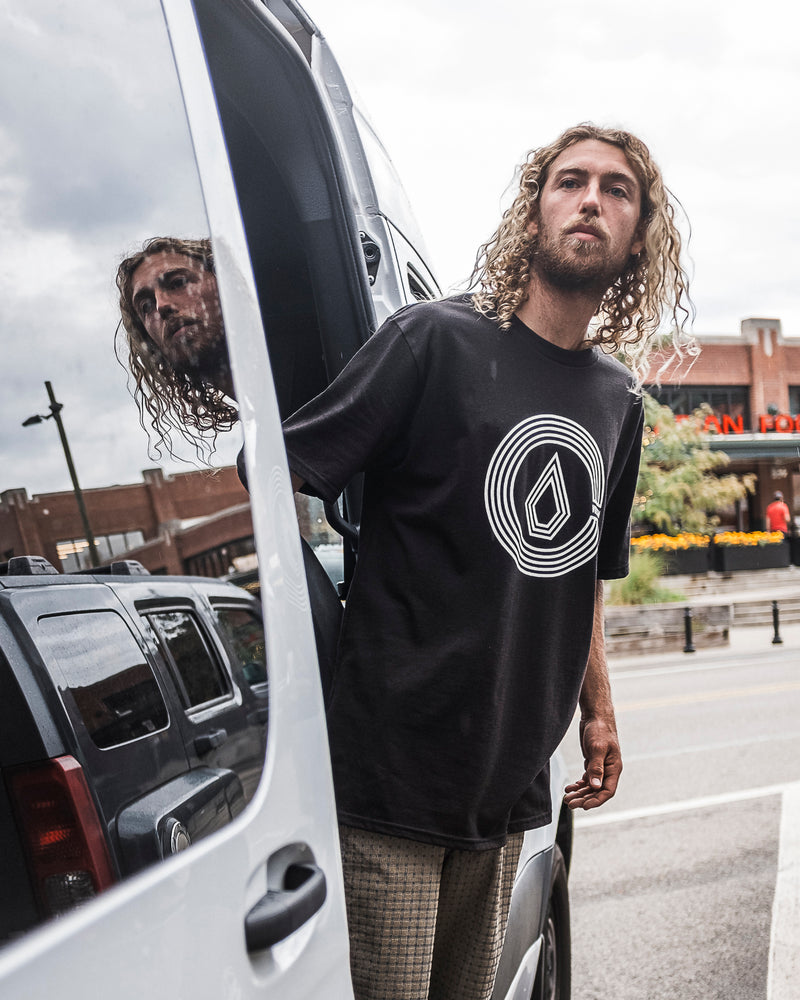 A man with long curly hair steps out of a white van, wearing the Volcom Men's Circle Split Short Sleeve Tee. His reflection shimmers in the window while buildings and flowers fill the background.