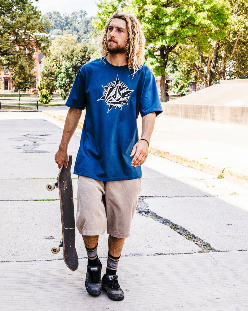 A young man with long curly hair walks outdoors holding a skateboard, wearing the Volcom Men's Yoink Short Sleeve Tee in blue, beige knee-length shorts, black socks, and sneakers. Trees and concrete structures are in the background.