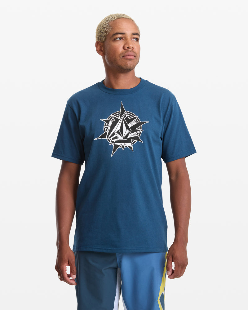 A person with short, blond hair wears the Volcom Men's Yoink Short Sleeve Tee in dark blue, showcasing a large white compass design, paired with blue shorts featuring a yellow stripe, standing against a plain white background.