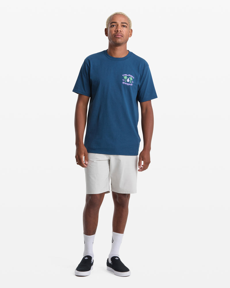 A person stands facing forward in the Volcom Men's Fed Up Short Sleeve Tee (dark blue, 100% cotton, classic fit), paired with light gray shorts, white socks, and black slip-on shoes against a plain white background.